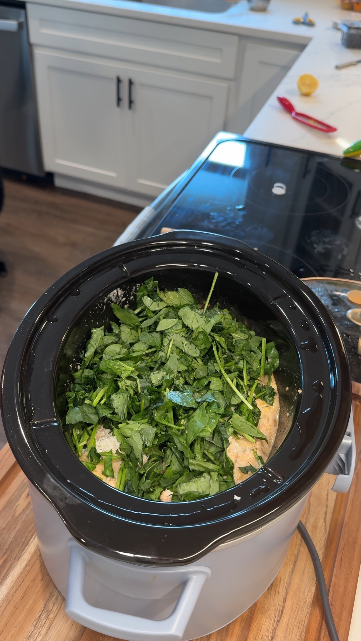 A slow cooker filled with chopped spinach and what appears to be uncooked chicken in a creamy sauce sits on a kitchen counter near a stove.
