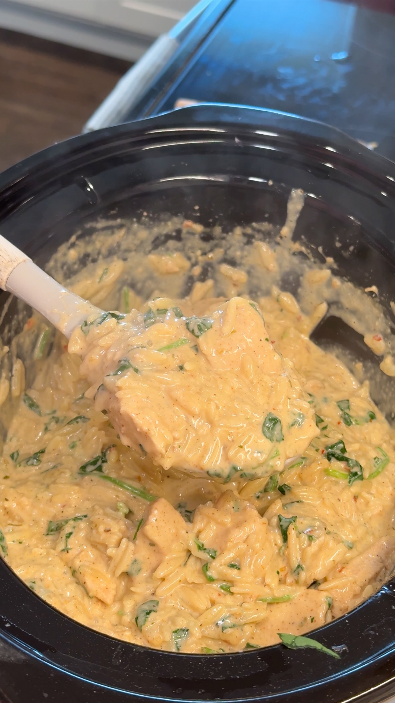 A close-up of a creamy, cheesy pasta dish with bits of spinach being scooped from a slow cooker with a white spatula.