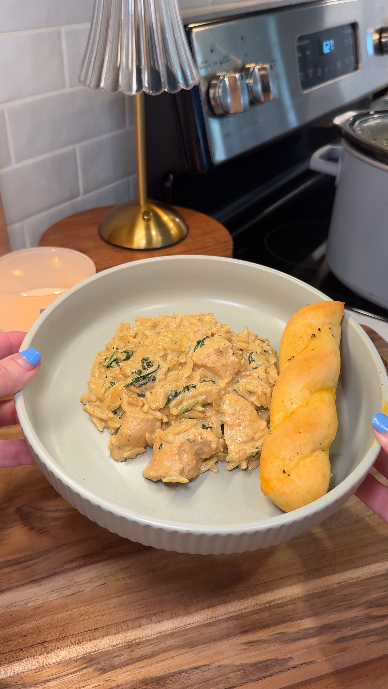A person with blue-painted nails holds a bowl of cowboy butter chicken orzo and a twisted breadstick. The background shows a stove, a lamp, and a candle on a wooden counter.