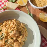 A bowl of creamy orzo pasta with chicken and spinach sits on a plate. Nearby are lemon wedges, raw orzo in a small bowl, spinach leaves, and pieces of Parmesan on a red checkered cloth.
