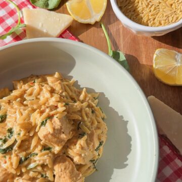 A bowl of creamy orzo pasta with chicken and spinach sits on a plate. Nearby are lemon wedges, raw orzo in a small bowl, spinach leaves, and pieces of Parmesan on a red checkered cloth.