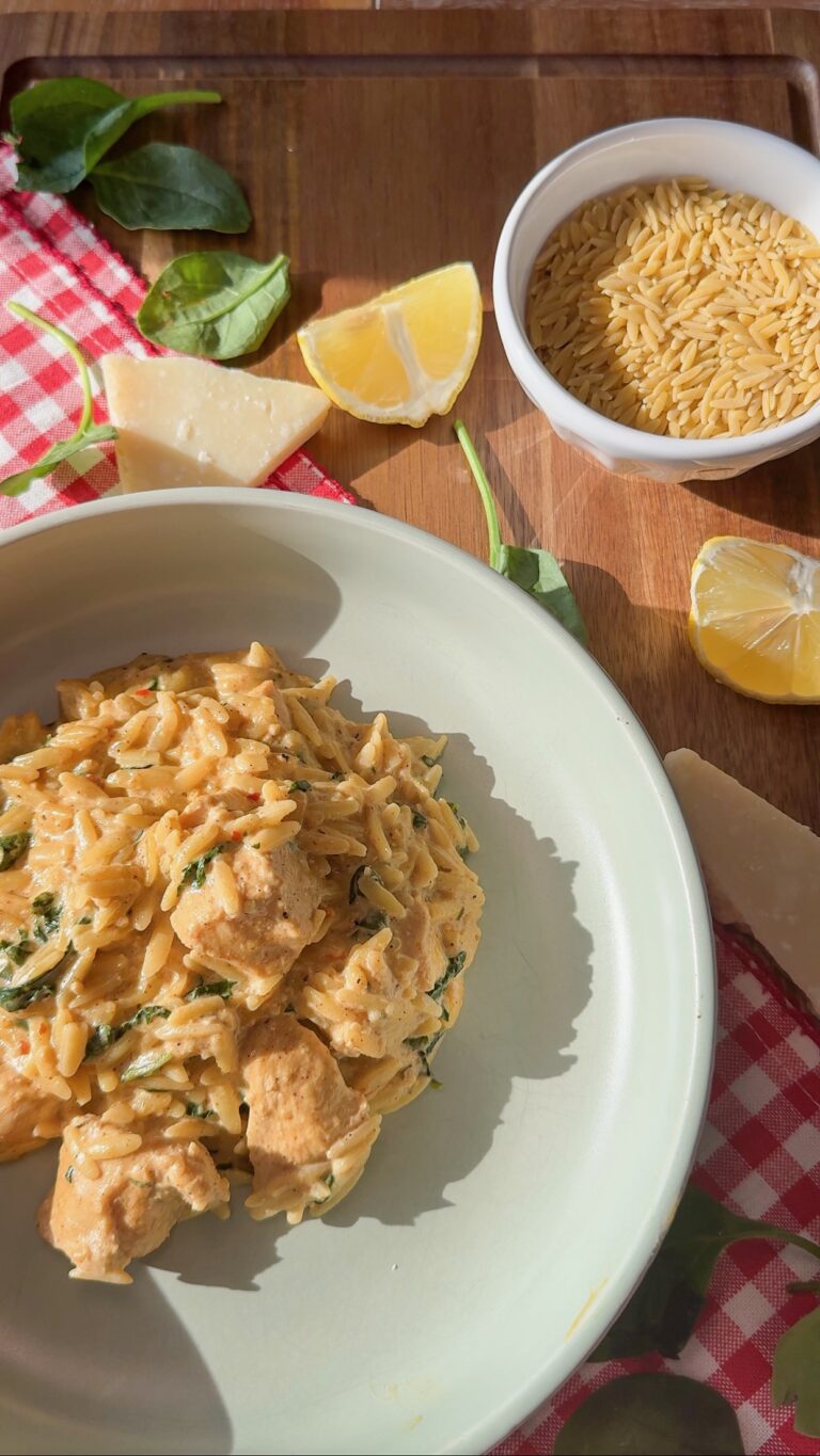 A bowl of creamy orzo pasta with chicken and spinach sits on a plate. Nearby are lemon wedges, raw orzo in a small bowl, spinach leaves, and pieces of Parmesan on a red checkered cloth.