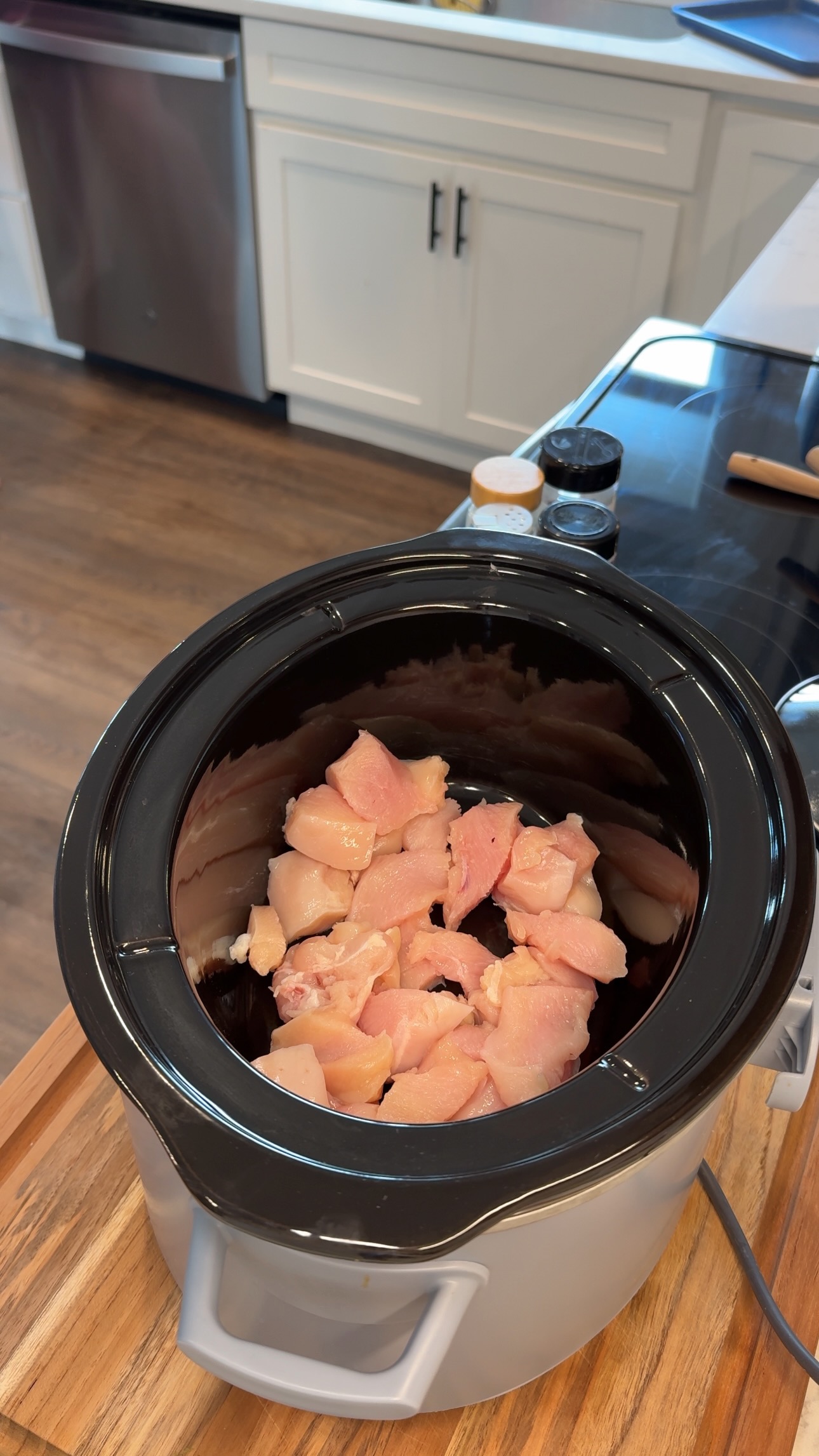 Raw, diced chicken pieces are placed inside a black slow cooker on a kitchen counter. The countertop has spice jars and a rolling pin nearby, and white cabinets and a dishwasher are visible in the background.