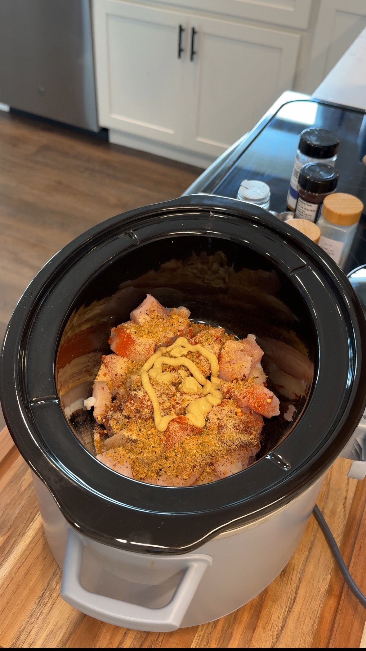A slow cooker with raw, seasoned chicken topped with a squiggle of mustard sits on a wooden countertop. Spice containers and kitchen cabinets are visible in the background.