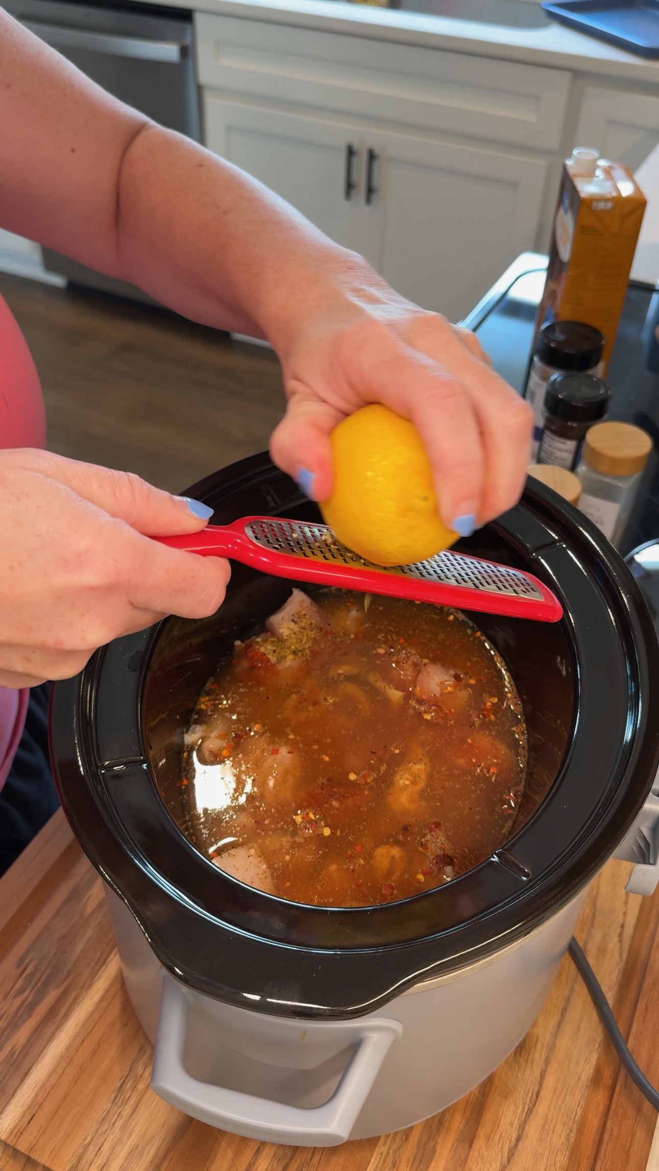A person grates a lemon over a slow cooker filled with seasoned liquid and meat. Various spices and ingredients are on the counter in a kitchen setting.