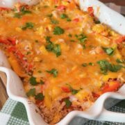 A baked casserole topped with melted cheese, chopped cilantro, and colorful bell peppers sits in a white dish on a green and white checkered cloth.