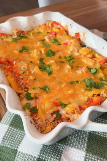A baked casserole topped with melted cheese, chopped cilantro, and colorful bell peppers sits in a white dish on a green and white checkered cloth.