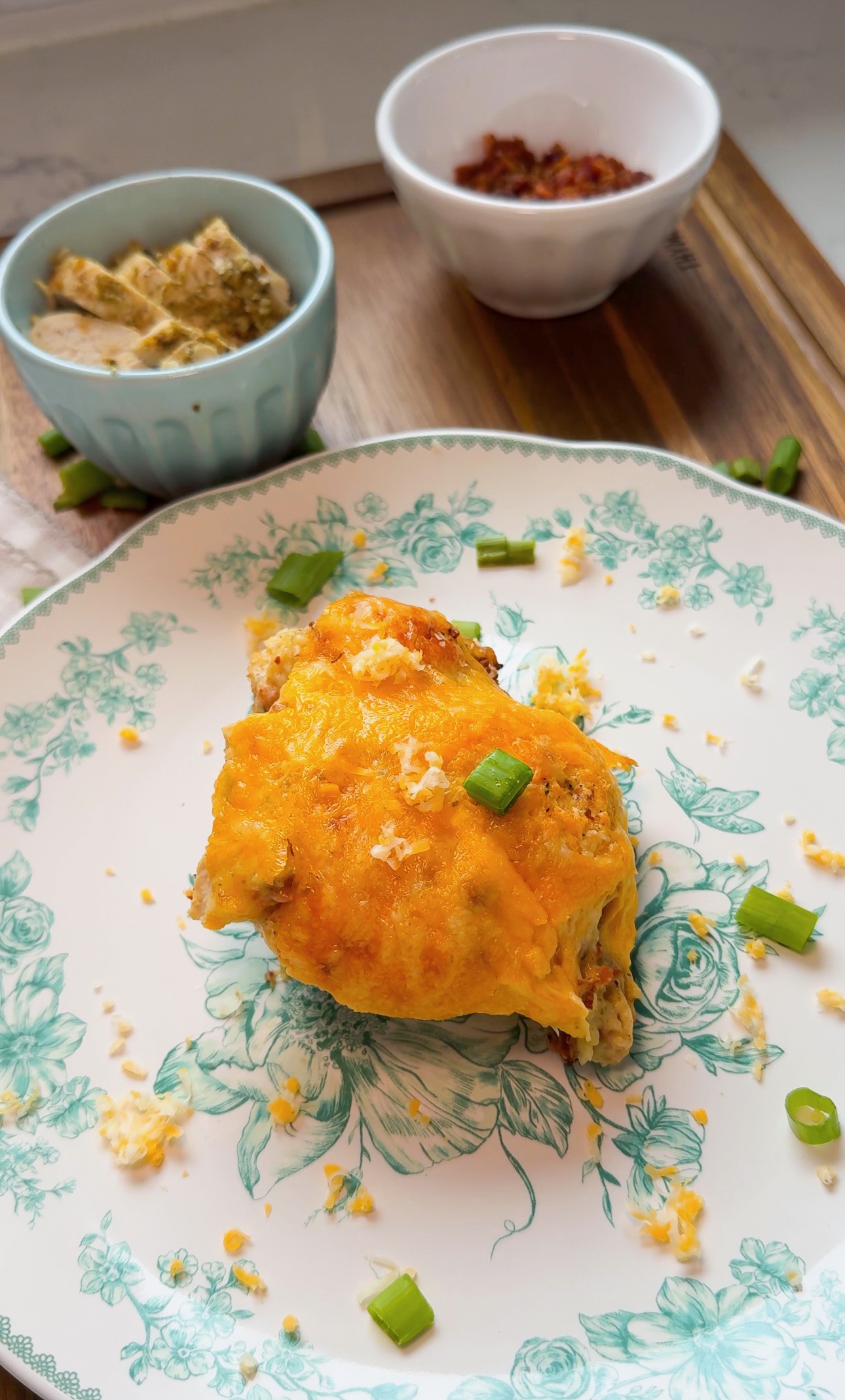A plate with a cheesy baked dish topped with chopped green onions, accompanied by small bowls of dip and diced toppings in the background on a wooden board.