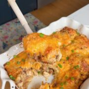 A hand holds a spatula lifting a cheesy baked casserole from a white dish. The casserole is topped with melted cheese and scattered green onions, with a portion already served.