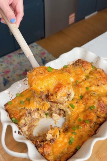 A hand holds a spatula lifting a cheesy baked casserole from a white dish. The casserole is topped with melted cheese and scattered green onions, with a portion already served.