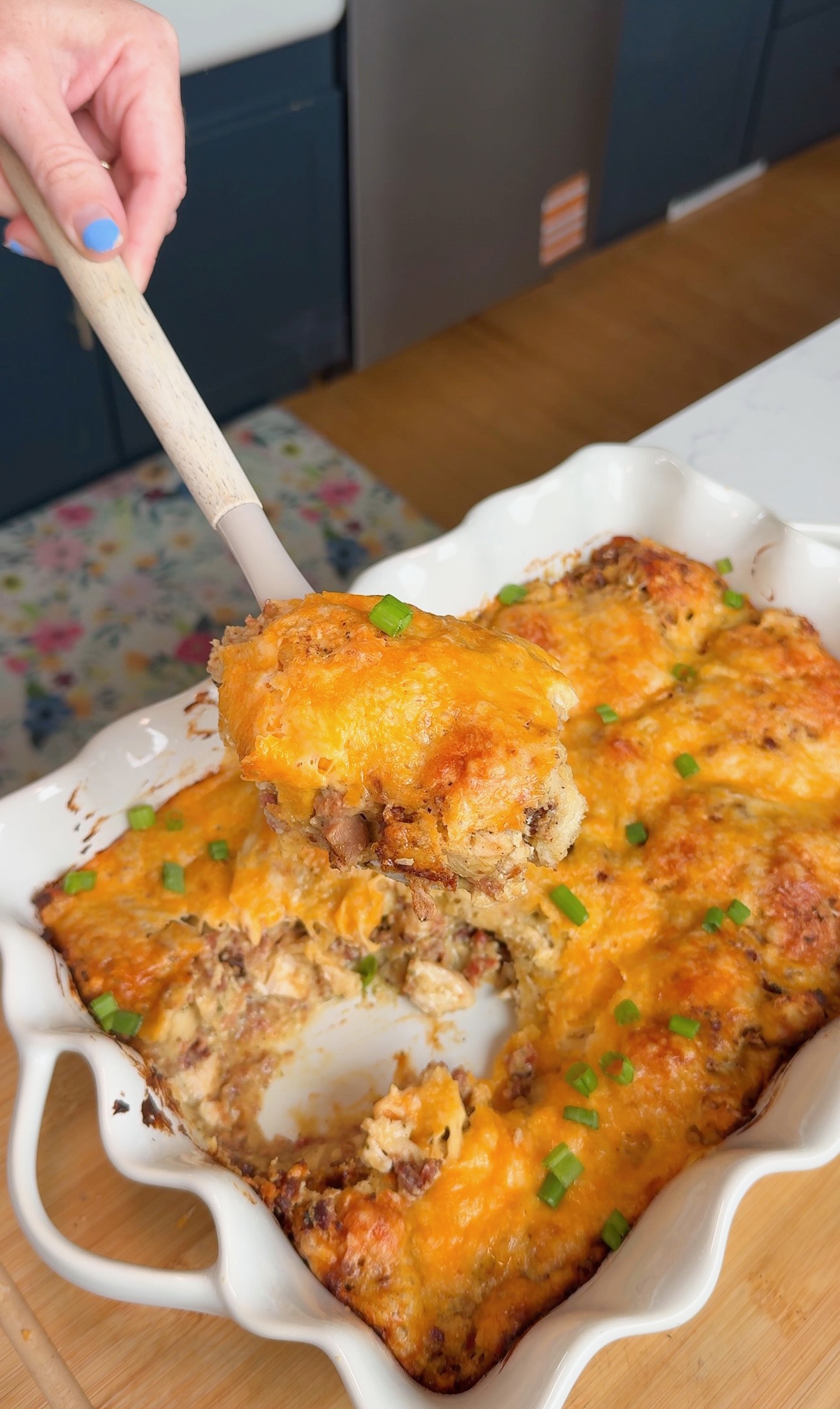 A hand holds a spatula lifting a cheesy baked casserole from a white dish. The casserole is topped with melted cheese and scattered green onions, with a portion already served.