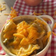 A small white dish of cheesy ravioli topped with shredded cheddar sits on a red checkered cloth. An onion and more cheddar cheese are in the background.