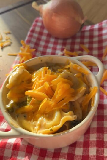 A small white dish of cheesy ravioli topped with shredded cheddar sits on a red checkered cloth. An onion and more cheddar cheese are in the background.