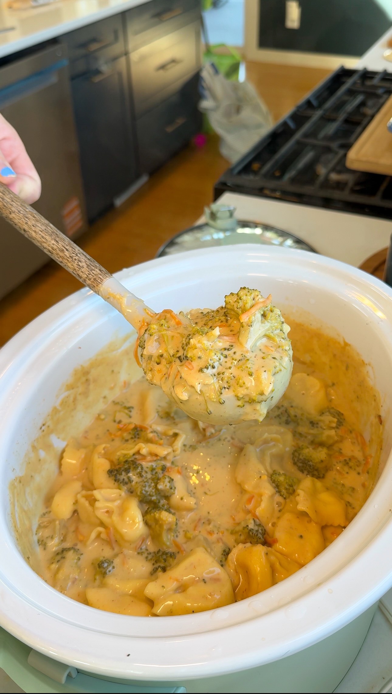 A wooden ladle scoops creamy chicken, broccoli, and carrots from a white slow cooker on a kitchen counter.