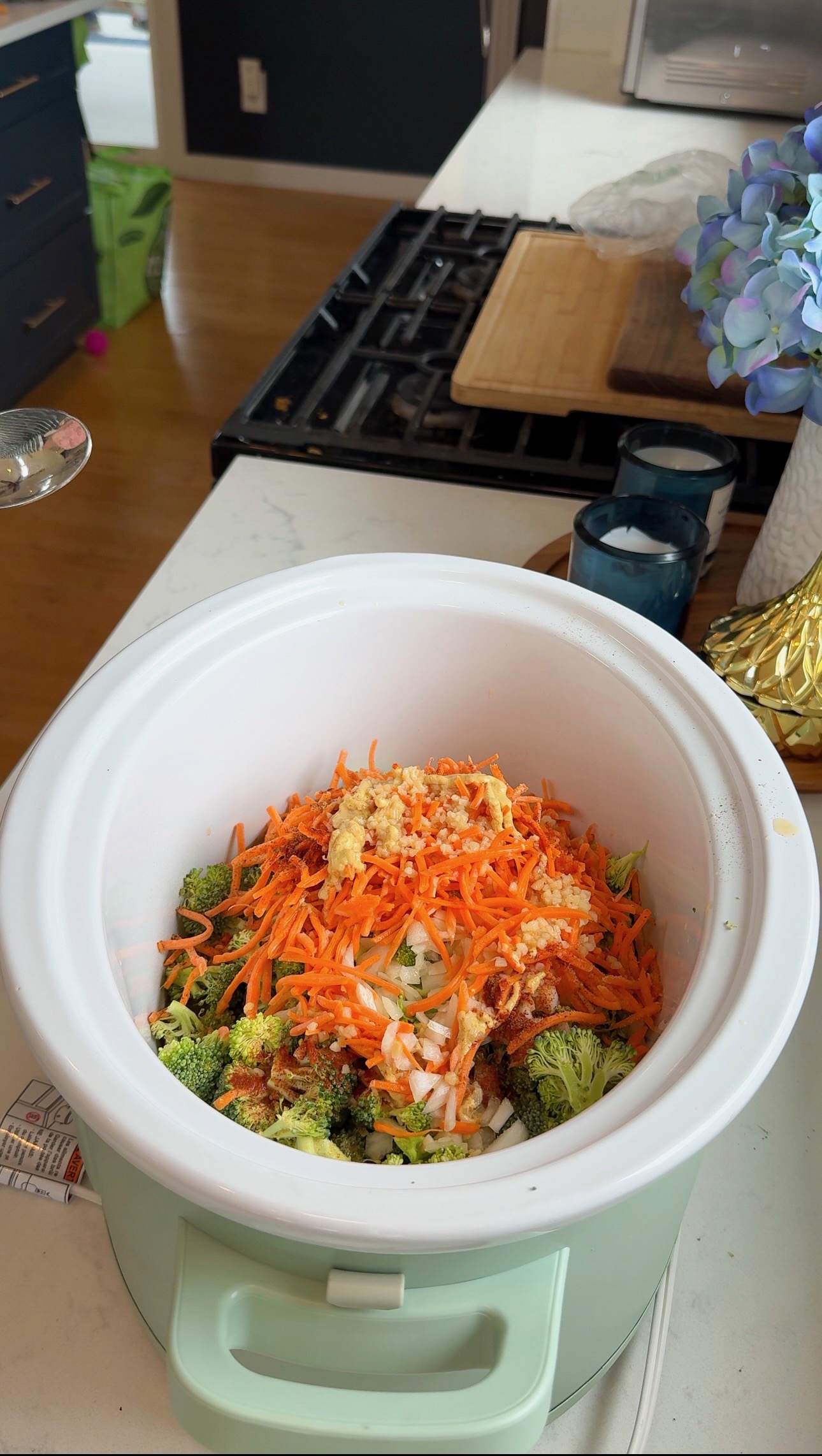A slow cooker on a kitchen counter filled with chopped broccoli, shredded carrots, onions, and seasonings, ready to be cooked.