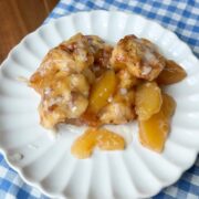 A serving of cinnamon rolls and peach slices with icing on a white plate, placed on a blue and white checkered cloth on a wooden table.