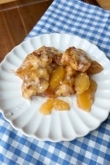 A serving of cinnamon rolls and peach slices with icing on a white plate, placed on a blue and white checkered cloth on a wooden table.