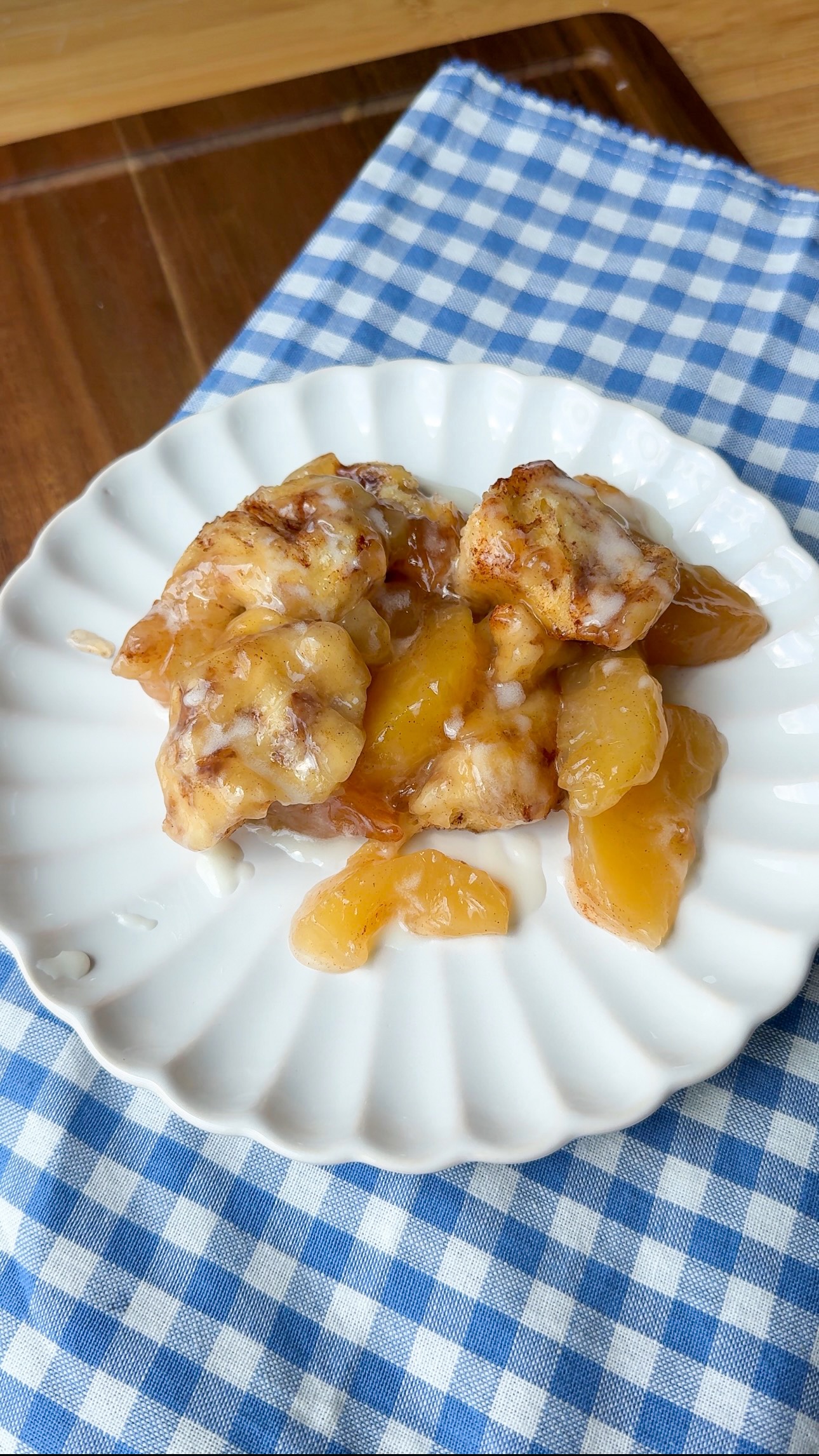 A serving of cinnamon rolls and peach slices with icing on a white plate, placed on a blue and white checkered cloth on a wooden table.