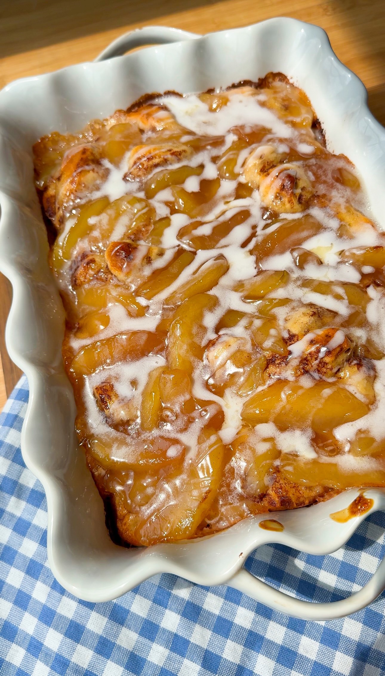 A baked dessert in a white scalloped dish, topped with golden apple slices and white icing, sits on a blue and white checkered cloth in sunlight.