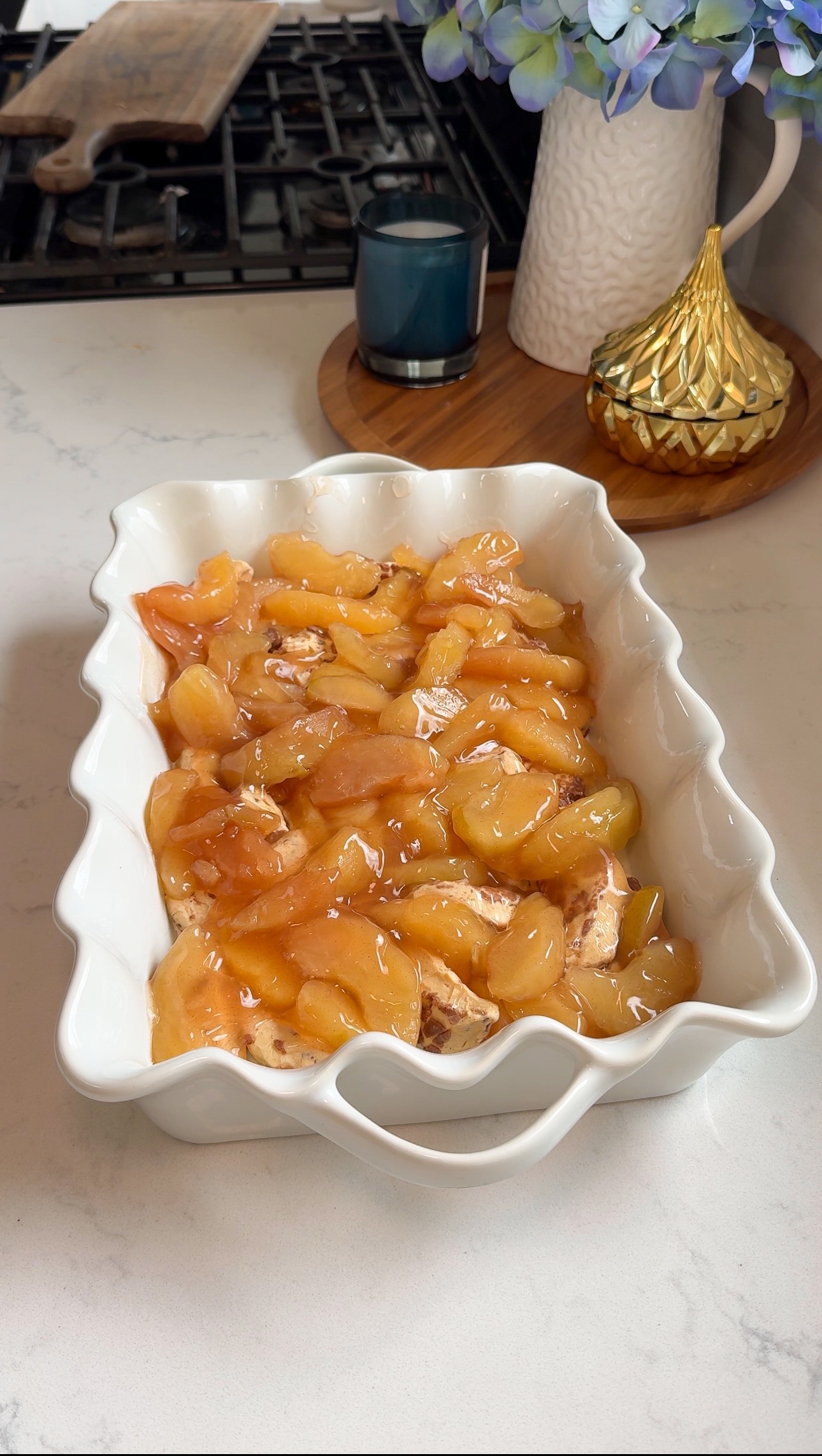 A white baking dish filled with sliced apples in syrup sits on a kitchen counter near a blue candle, decorative jar, and vase of flowers, with a stove in the background.