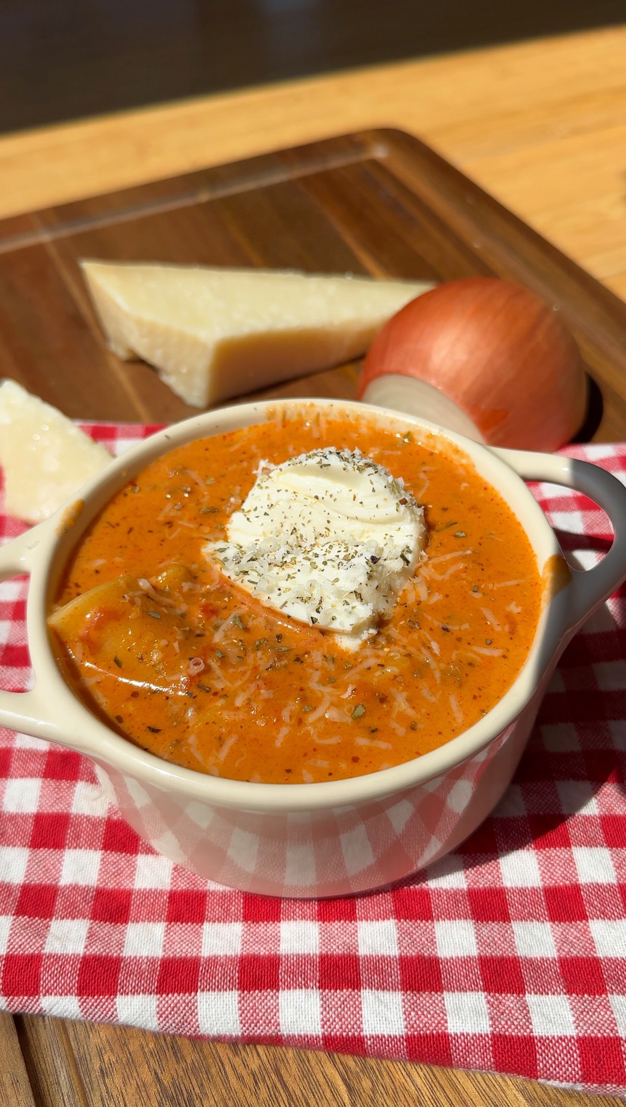 A bowl of creamy tomato soup topped with herbs and sour cream sits on a red and white checkered cloth, with a wedge of Parmesan cheese and an onion on a wooden surface nearby.