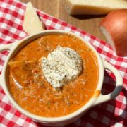 A bowl of creamy tomato soup topped with herbs and sour cream sits on a red and white checkered cloth, with a wedge of Parmesan cheese and an onion on a wooden surface nearby.