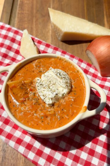 A bowl of creamy tomato soup topped with herbs and sour cream sits on a red and white checkered cloth, with a wedge of Parmesan cheese and an onion on a wooden surface nearby.