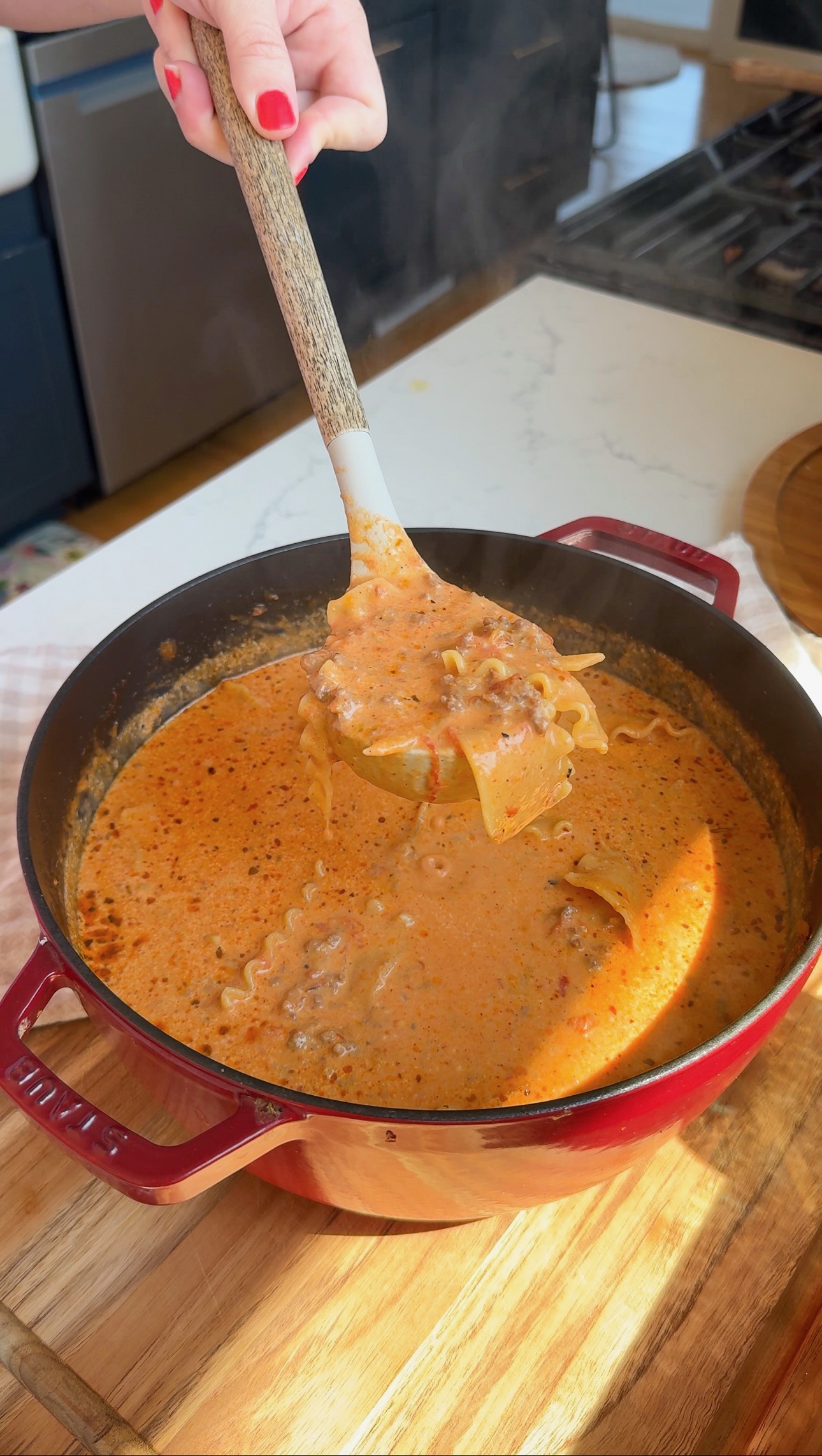 A hand holds a ladle scooping creamy lasagna soup with noodles and ground meat from a red pot on a wooden countertop. Steam rises from the soup.