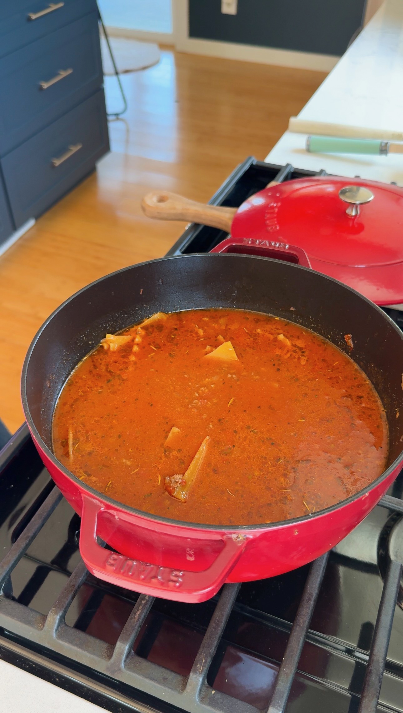 A red Dutch oven on a stove contains a rich, red broth with visible herbs and pieces of pasta or vegetables. A matching red lid rests nearby on the stovetop. Bright kitchen with wooden floors in the background.