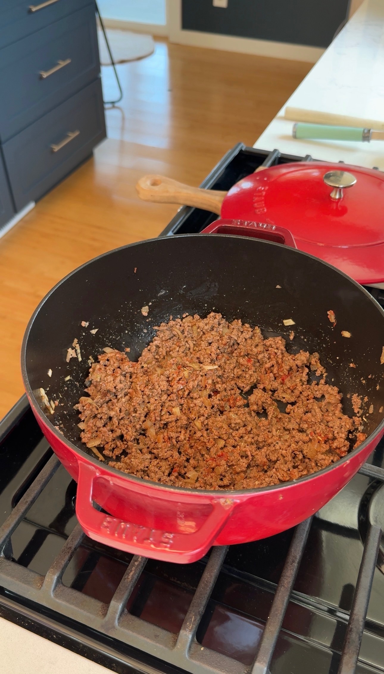 Ground meat with onions and spices is cooking in a red Dutch oven on a stove, with a matching red lid and wooden spoon nearby. The kitchen features wood floors and dark cabinets.