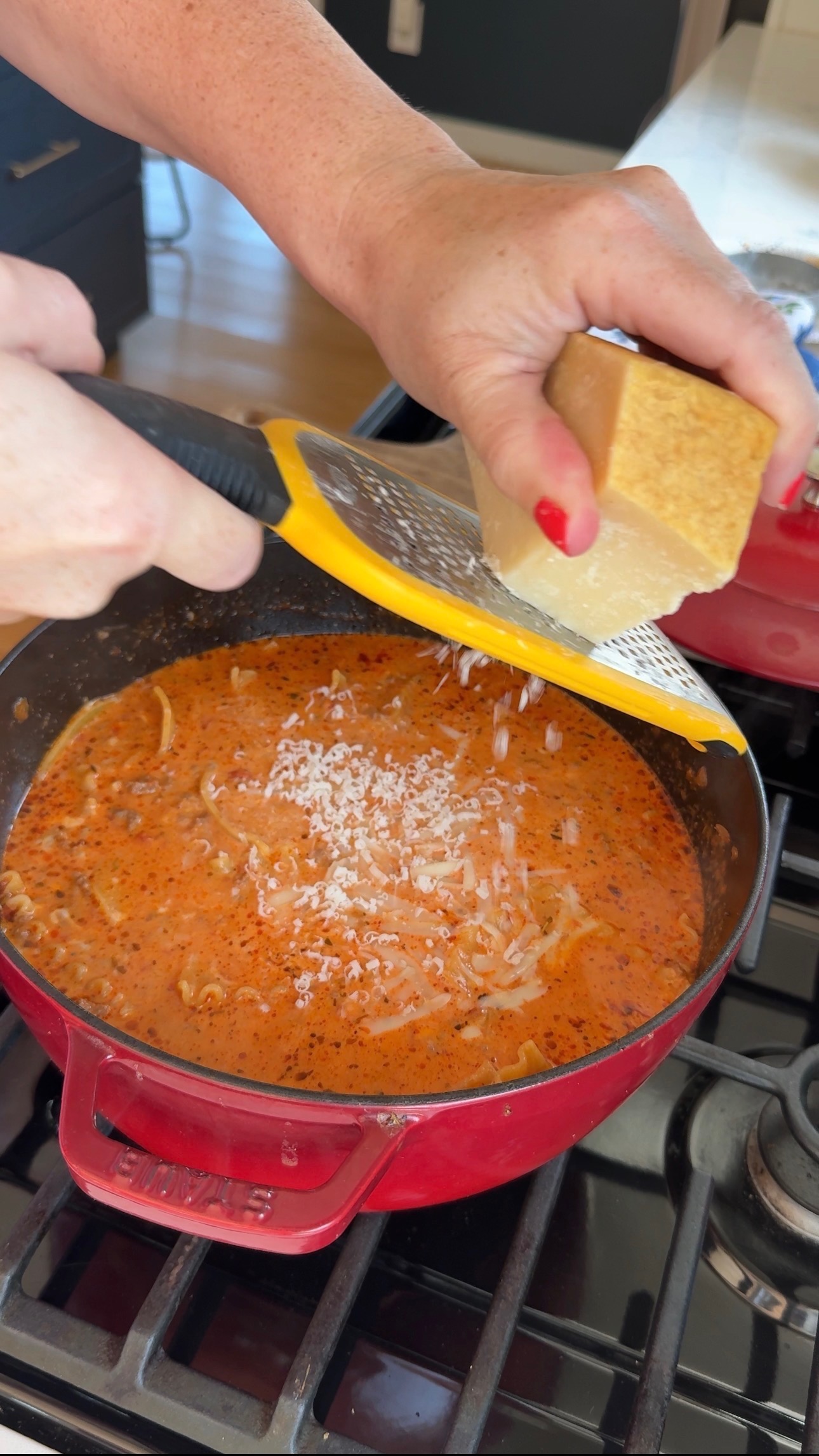 A hand grates cheese over a pot of creamy tomato soup on a stove. The soup is in a red Dutch oven, and the person has red nail polish.