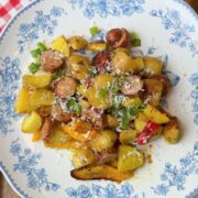A floral plate holds roasted potatoes, sausage slices, and colorful bell peppers, topped with grated cheese. The plate sits on a red gingham napkin with green onions and cheese wedges nearby on a wooden table.
