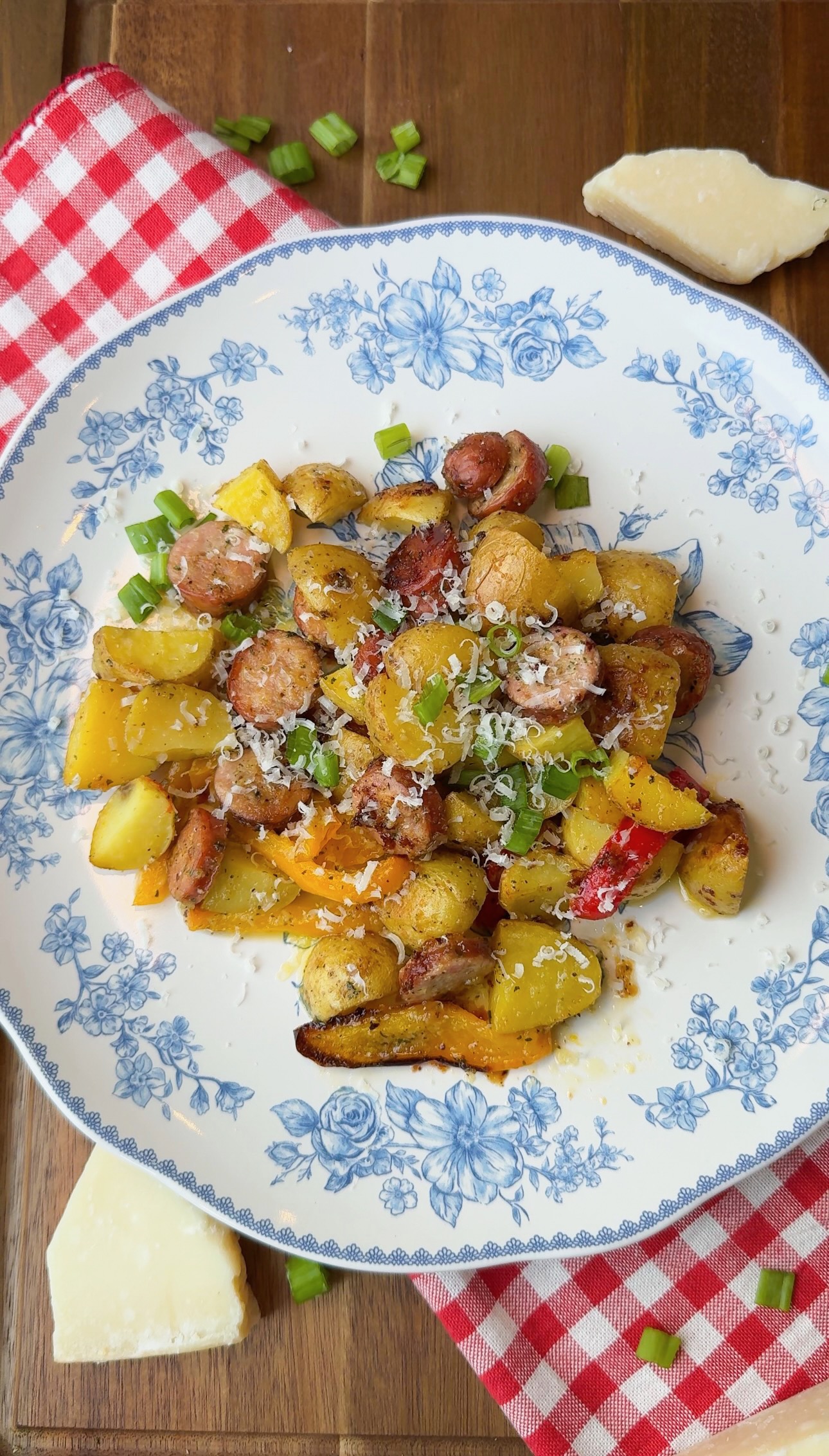A floral plate holds roasted potatoes, sausage slices, and colorful bell peppers, topped with grated cheese. The plate sits on a red gingham napkin with green onions and cheese wedges nearby on a wooden table.
