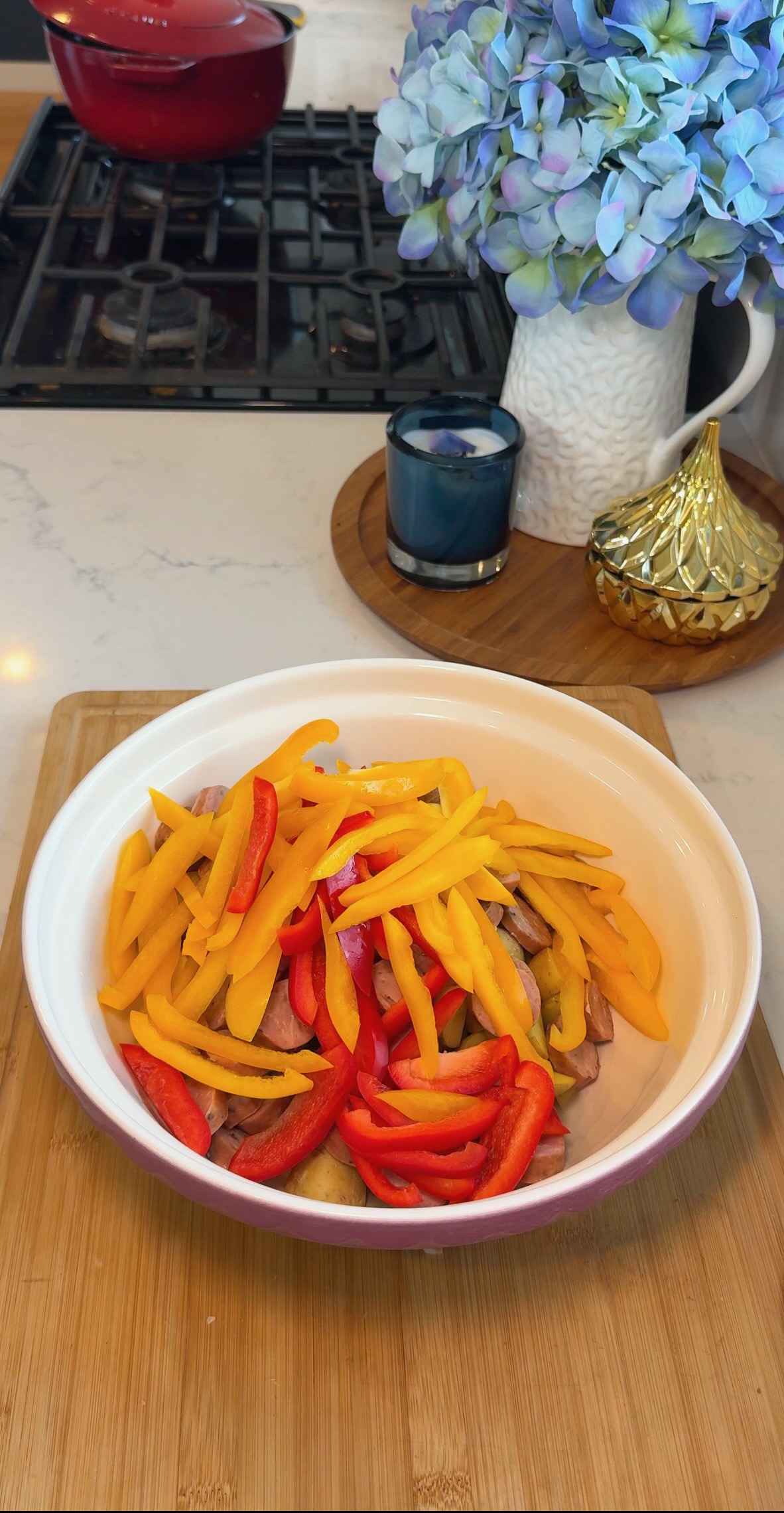 A white bowl with sliced red and yellow bell peppers sits on a wooden cutting board in a kitchen. Behind it are a blue candle, a gold decorative item, blue flowers, and a stove with a red pot.