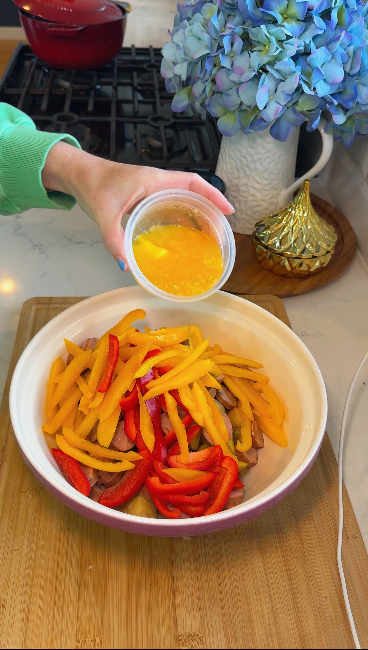 A hand pours a small cup of yellow liquid over a bowl of sliced red and yellow bell peppers on a kitchen counter. A blue hydrangea and gold container are in the background.