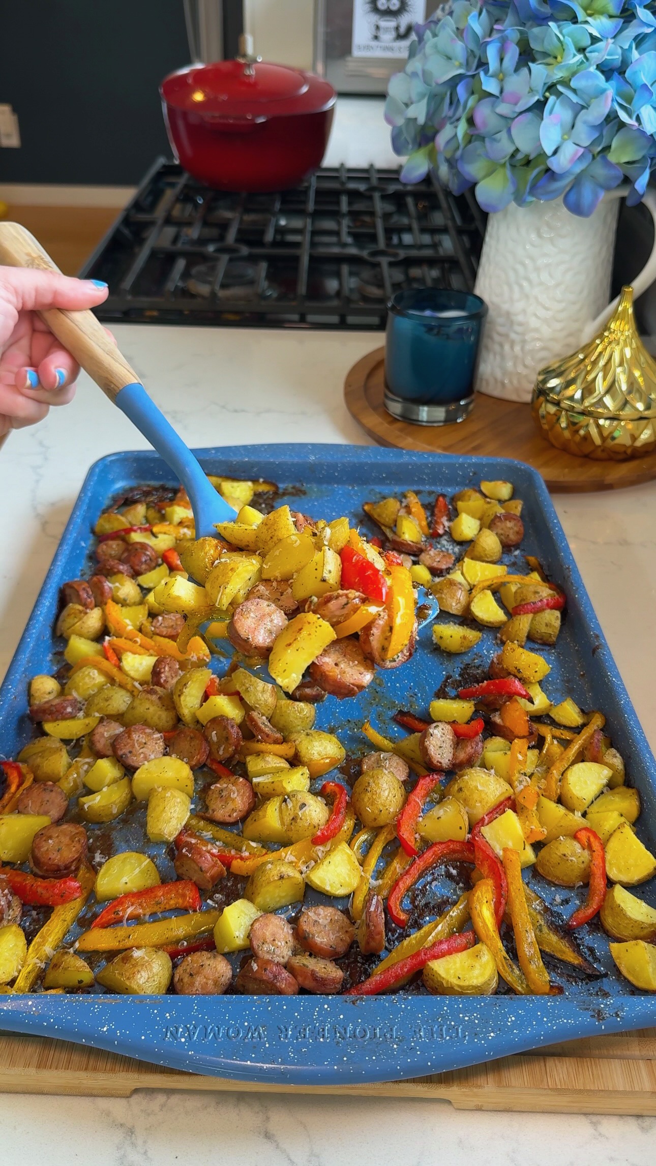 A hand serves roasted potatoes, sausage, and bell peppers from a blue baking tray on a kitchen counter with flowers, a candle, and decor in the background.