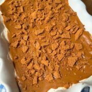A white scalloped dish holds a dessert topped with a caramel-colored layer and crumbled cookies, sitting on a blue and white checkered cloth on a wooden surface.