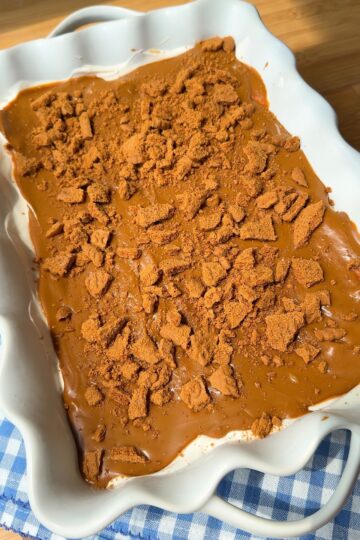 A white scalloped dish holds a dessert topped with a caramel-colored layer and crumbled cookies, sitting on a blue and white checkered cloth on a wooden surface.