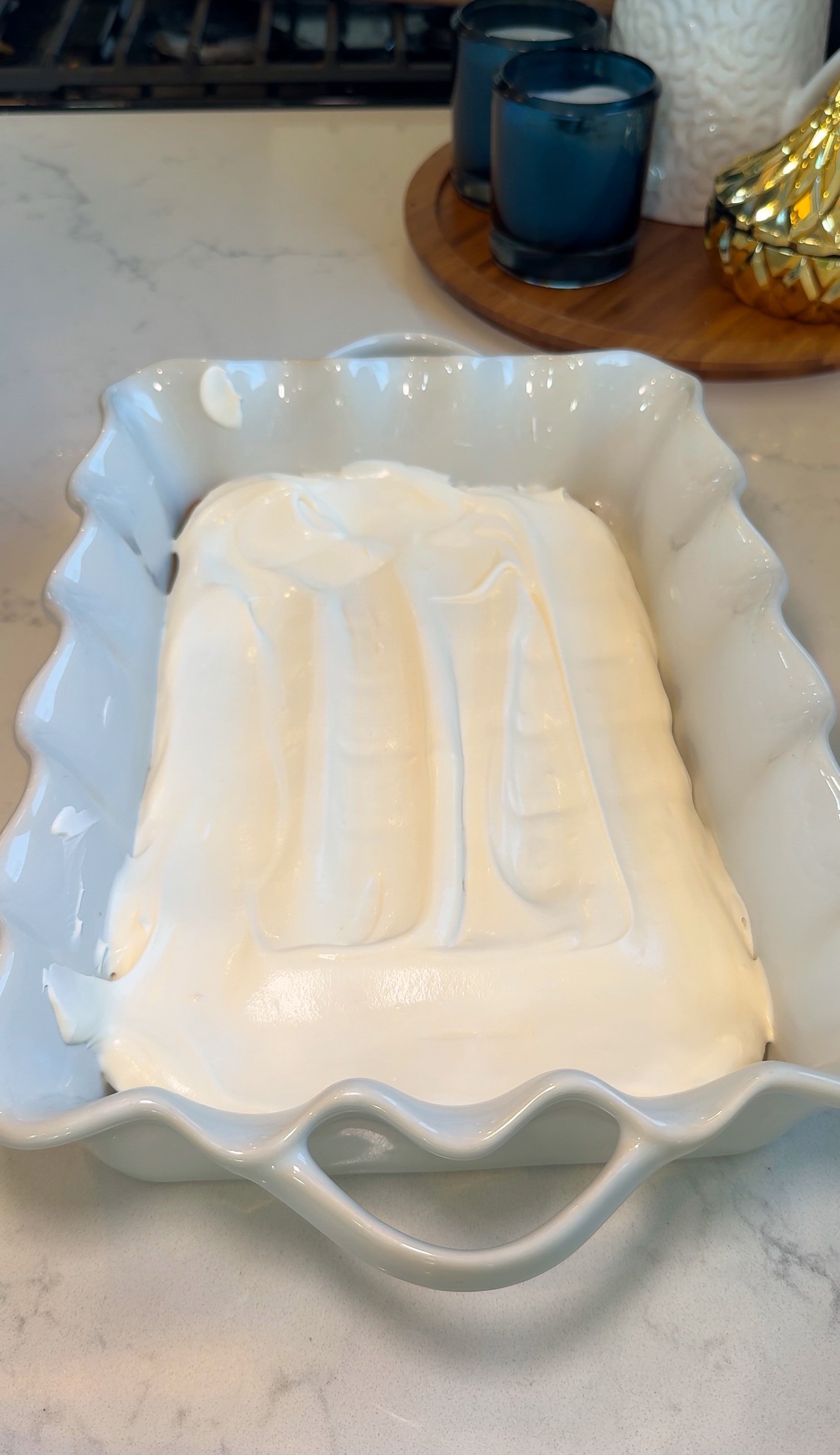 A white ceramic dish filled with a smooth layer of white whipped cream or frosting sits on a countertop, with candles and decor in the background.