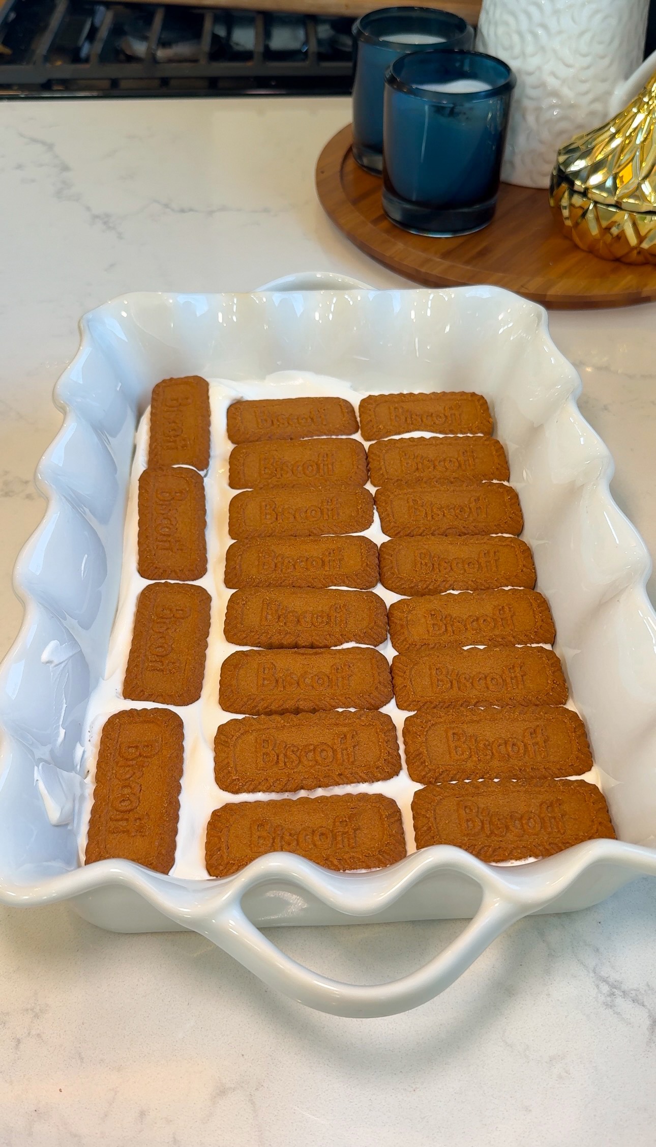 A white baking dish with a layer of Biscoff cookies arranged neatly over a layer of white cream, on a kitchen counter with candles and decor in the background.