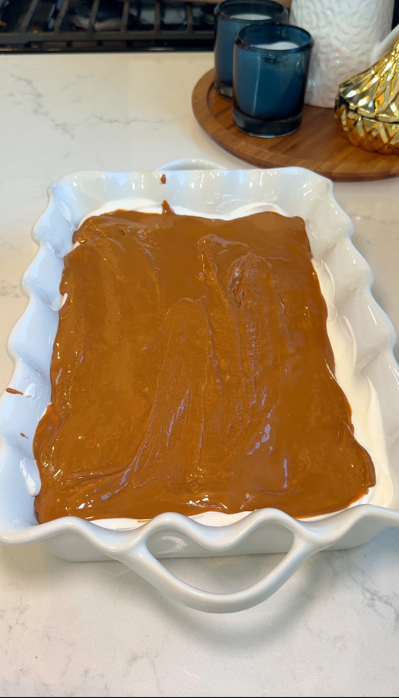 A white ceramic baking dish with wavy edges is filled with a smooth, caramel-brown mixture. The dish sits on a light countertop near blue candles and a gold decorative object.