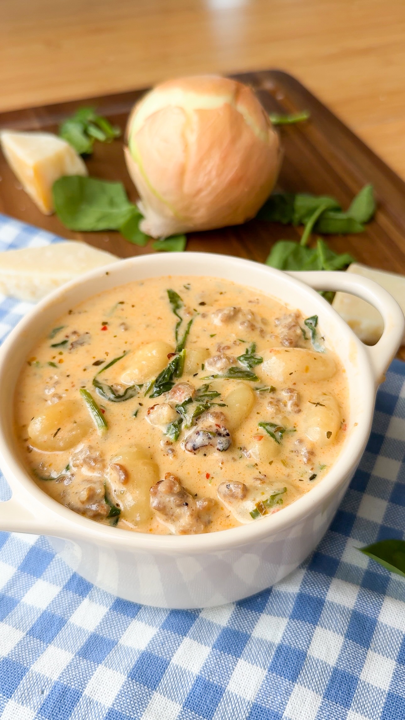 Creamy gnocchi soup with sausage and spinach in a white bowl, placed on a blue checkered cloth. In the background, a cutting board holds a peeled onion, spinach leaves, and a lemon wedge.