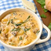 A white bowl of creamy soup with sausage, gnocchi, and spinach sits on a blue checkered cloth, with cheese and greens on a wooden board nearby.
