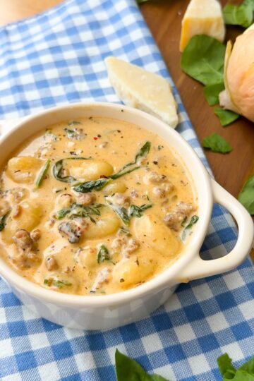 A white bowl of creamy soup with sausage, gnocchi, and spinach sits on a blue checkered cloth, with cheese and greens on a wooden board nearby.