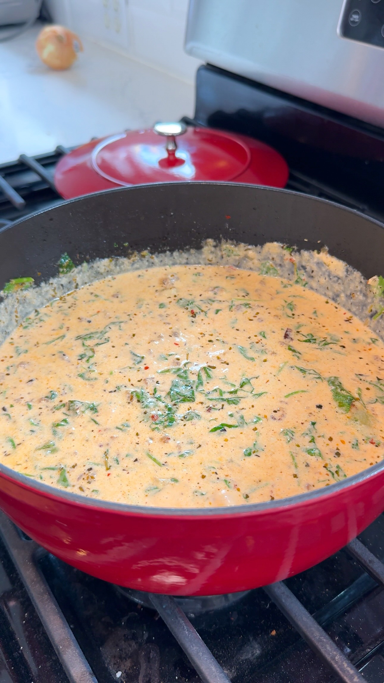 A red pot on a stove filled with a creamy, orange-colored soup or sauce, mixed with green leafy herbs and spices. A red pot lid is in the background.