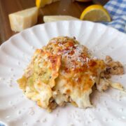 A serving of cheesy lasagna sits on a white plate, topped with grated cheese. In the background are a whole onion, lemon wedges, a cheese wedge, and a blue checkered cloth.