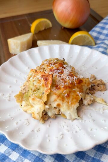 A serving of cheesy lasagna sits on a white plate, topped with grated cheese. In the background are a whole onion, lemon wedges, a cheese wedge, and a blue checkered cloth.