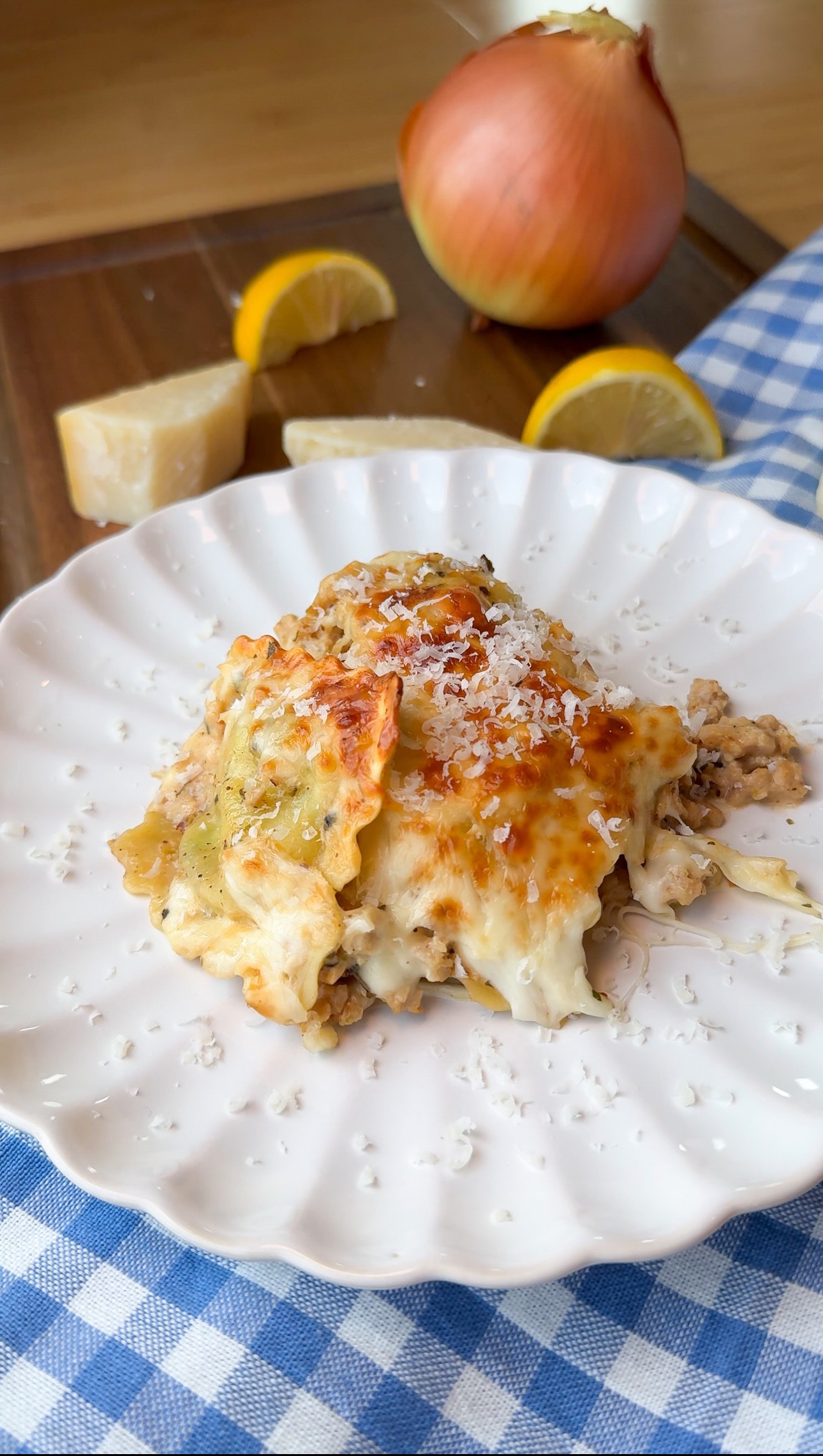 A serving of cheesy lasagna sits on a white plate, topped with grated cheese. In the background are a whole onion, lemon wedges, a cheese wedge, and a blue checkered cloth.