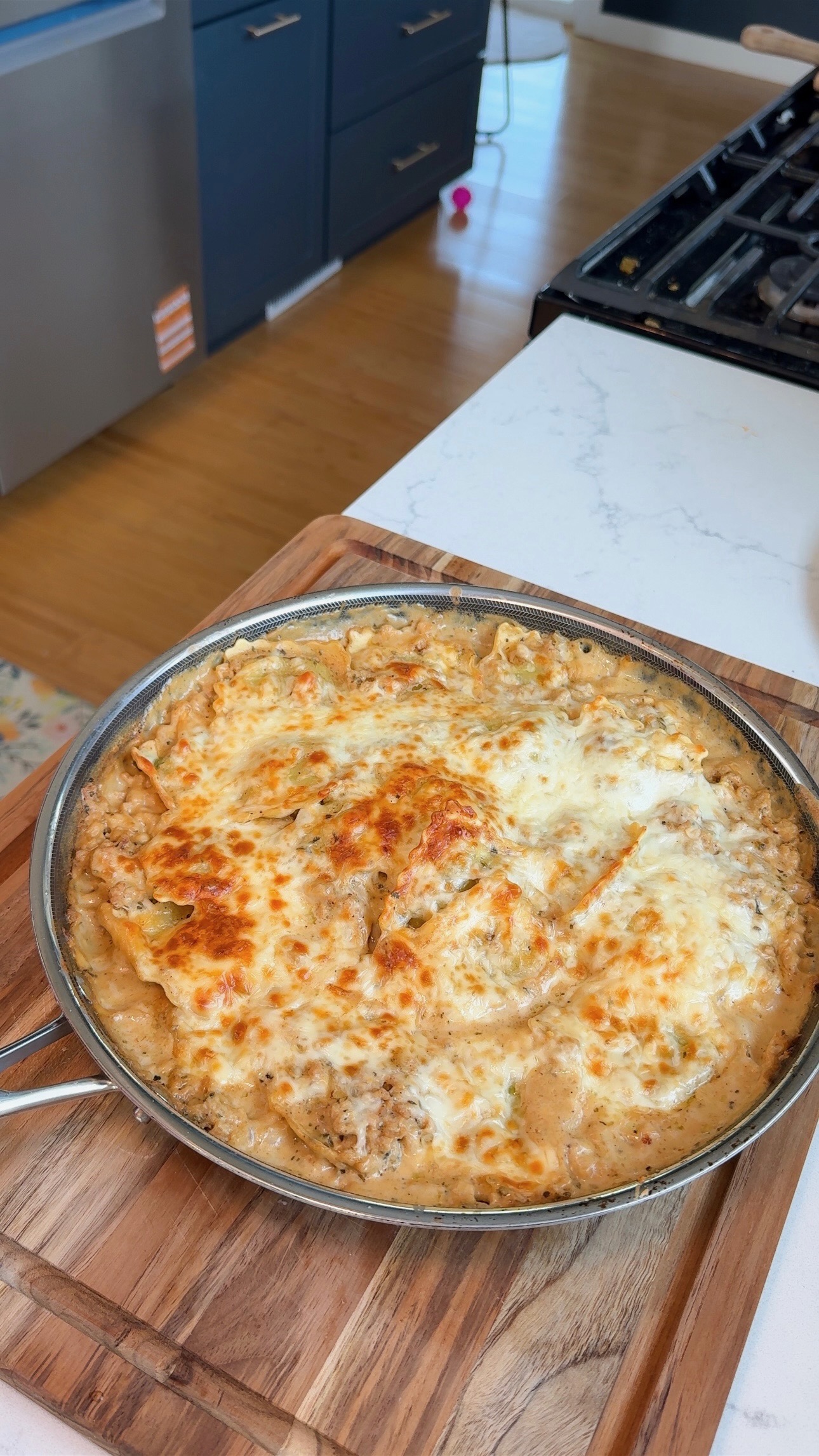 A skillet of baked cheesy casserole on a wooden board, with melted golden-brown cheese on top, sits on a white kitchen counter near a stove.