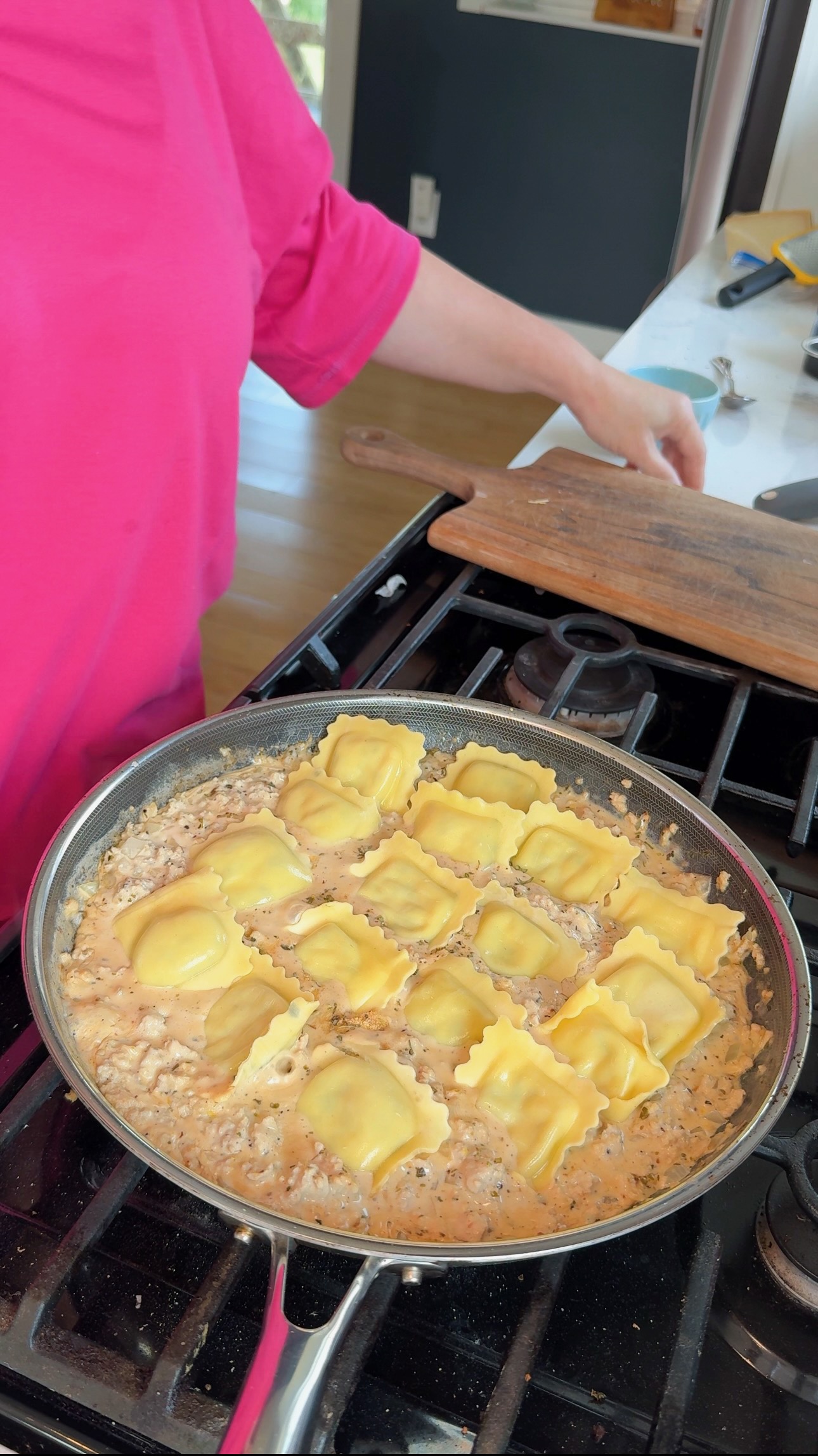 A person in a pink shirt stands near a stove where ravioli cooks in a creamy sauce in a pan. The counter has a wooden board and utensils.